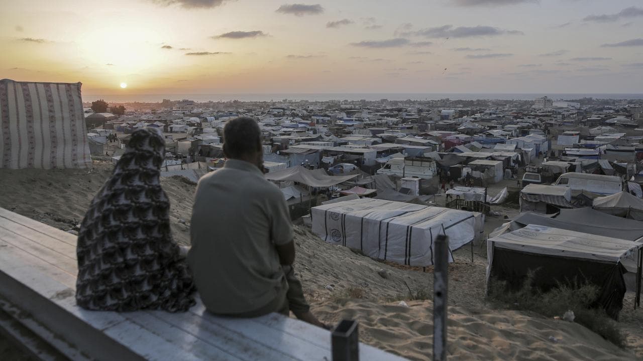 Two people sit on a wall and look over a tent camp in Gaza.