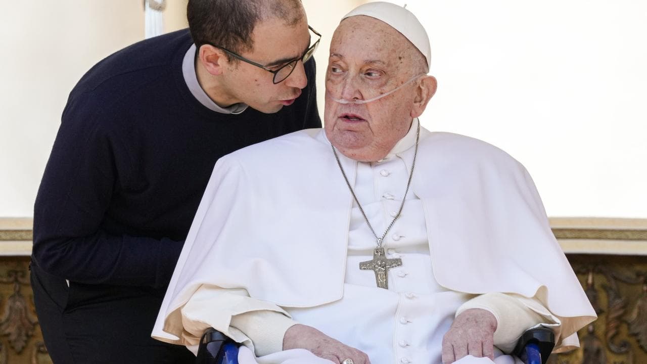 Pope Francis in a wheelchair listens to an aide