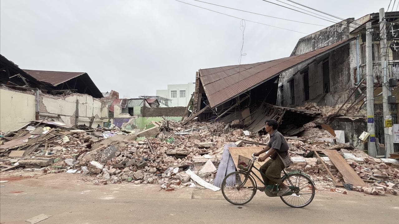 A man rides a bike past a damaged building